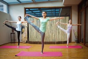 Three people are performing yoga poses on pink mats in a serene indoor space with wooden floors and a bamboo backdrop. They are balancing on one leg while holding the other leg up near head level, with arms extended. The atmosphere is calm and focused.