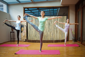 Three people are performing yoga poses on pink mats in a serene indoor space with wooden floors and a bamboo backdrop. They are balancing on one leg while holding the other leg up near head level, with arms extended. The atmosphere is calm and focused.