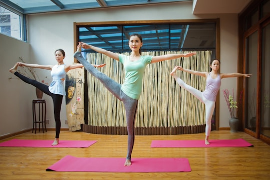 Three people are performing yoga poses on pink mats in a serene indoor space with wooden floors and a bamboo backdrop. They are balancing on one leg while holding the other leg up near head level, with arms extended. The atmosphere is calm and focused.
