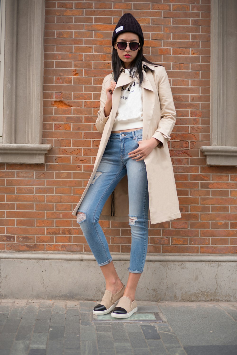 woman in white coat and blue denim jeans leaning on brown brick wall
