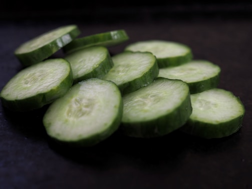 Slices of cucumbers are arranged in a slightly overlapping pattern on a dark surface. The cucumbers are evenly cut and display a crisp, fresh texture with a light green interior and darker green skin.