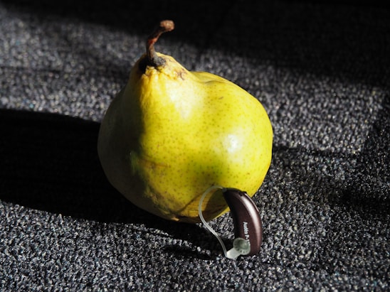 A yellow pear with speckles is placed on a textured dark surface. Attached to the pear is a small hearing aid device labeled 'Audio P'. The lighting creates distinct shadows and highlights across the pear and the surface.