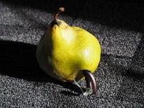 A yellow pear with speckles is placed on a textured dark surface. Attached to the pear is a small hearing aid device labeled 'Audio P'. The lighting creates distinct shadows and highlights across the pear and the surface.