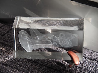 A transparent glass block featuring an etched design of the inner ear anatomy, displaying detailed structures like the cochlea. It is positioned on a textured, dark gray surface with a hearing aid resting partially inside the block.