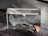 A transparent glass block featuring an etched design of the inner ear anatomy, displaying detailed structures like the cochlea. It is positioned on a textured, dark gray surface with a hearing aid resting partially inside the block.