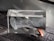 A transparent glass block featuring an etched design of the inner ear anatomy, displaying detailed structures like the cochlea. It is positioned on a textured, dark gray surface with a hearing aid resting partially inside the block.