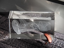 A transparent glass block featuring an etched design of the inner ear anatomy, displaying detailed structures like the cochlea. It is positioned on a textured, dark gray surface with a hearing aid resting partially inside the block.