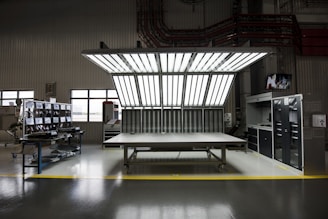 A large, well-lit workspace with a central table beneath a bright overhead lighting structure. The surroundings include a series of windows and various cabinets and desks, possibly indicating a laboratory or studio environment. Equipment and chairs are positioned around the periphery.