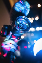 A hand holding a camera with a prominent lens, illuminated by vibrant blue and red lights. The background features a bokeh effect with various light spots creating an abstract ambiance.