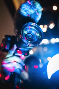 A hand holding a camera with a prominent lens, illuminated by vibrant blue and red lights. The background features a bokeh effect with various light spots creating an abstract ambiance.