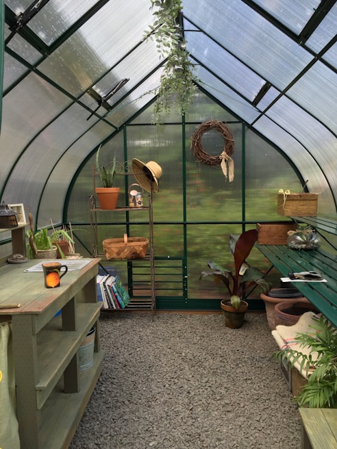 A quaint greenhouse interior filled with greenery, featuring a potting bench, a hanging plant, and gardening tools. Shelves hold books and pots, while a straw hat hangs beside a woven wreath on the wall. Sunlight filters through the translucent walls, adding a serene ambience.
