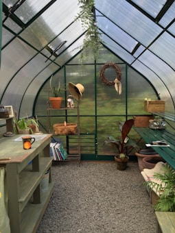 A quaint greenhouse interior filled with greenery, featuring a potting bench, a hanging plant, and gardening tools. Shelves hold books and pots, while a straw hat hangs beside a woven wreath on the wall. Sunlight filters through the translucent walls, adding a serene ambience.