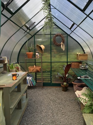 A quaint greenhouse interior filled with greenery, featuring a potting bench, a hanging plant, and gardening tools. Shelves hold books and pots, while a straw hat hangs beside a woven wreath on the wall. Sunlight filters through the translucent walls, adding a serene ambience.
