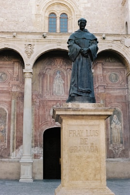 A statue of a man in robes stands atop a pedestal bearing the inscription 'Fray Luis de Granada'. The background features a stone facade with arches and detailed frescoes depicting religious figures. The scene exudes a sense of historical reverence and artistic detail.