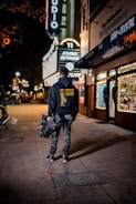 A person with a professional camera is walking along a dimly lit street at night. The person is wearing casual clothes including a hoodie with text on the back and camouflage pants. Neon signs, including one that reads 'STUDIO' and 'SUSHI COCKTAIL,' are visible, adding bright illumination. The area appears to be a commercial district with gift shops and restaurants.