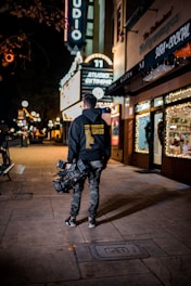 A person with a professional camera is walking along a dimly lit street at night. The person is wearing casual clothes including a hoodie with text on the back and camouflage pants. Neon signs, including one that reads 'STUDIO' and 'SUSHI COCKTAIL,' are visible, adding bright illumination. The area appears to be a commercial district with gift shops and restaurants.