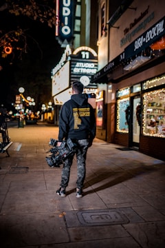 A person with a professional camera is walking along a dimly lit street at night. The person is wearing casual clothes including a hoodie with text on the back and camouflage pants. Neon signs, including one that reads 'STUDIO' and 'SUSHI COCKTAIL,' are visible, adding bright illumination. The area appears to be a commercial district with gift shops and restaurants.