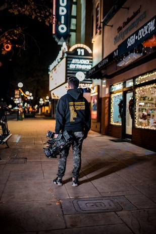 A person with a professional camera is walking along a dimly lit street at night. The person is wearing casual clothes including a hoodie with text on the back and camouflage pants. Neon signs, including one that reads 'STUDIO' and 'SUSHI COCKTAIL,' are visible, adding bright illumination. The area appears to be a commercial district with gift shops and restaurants.