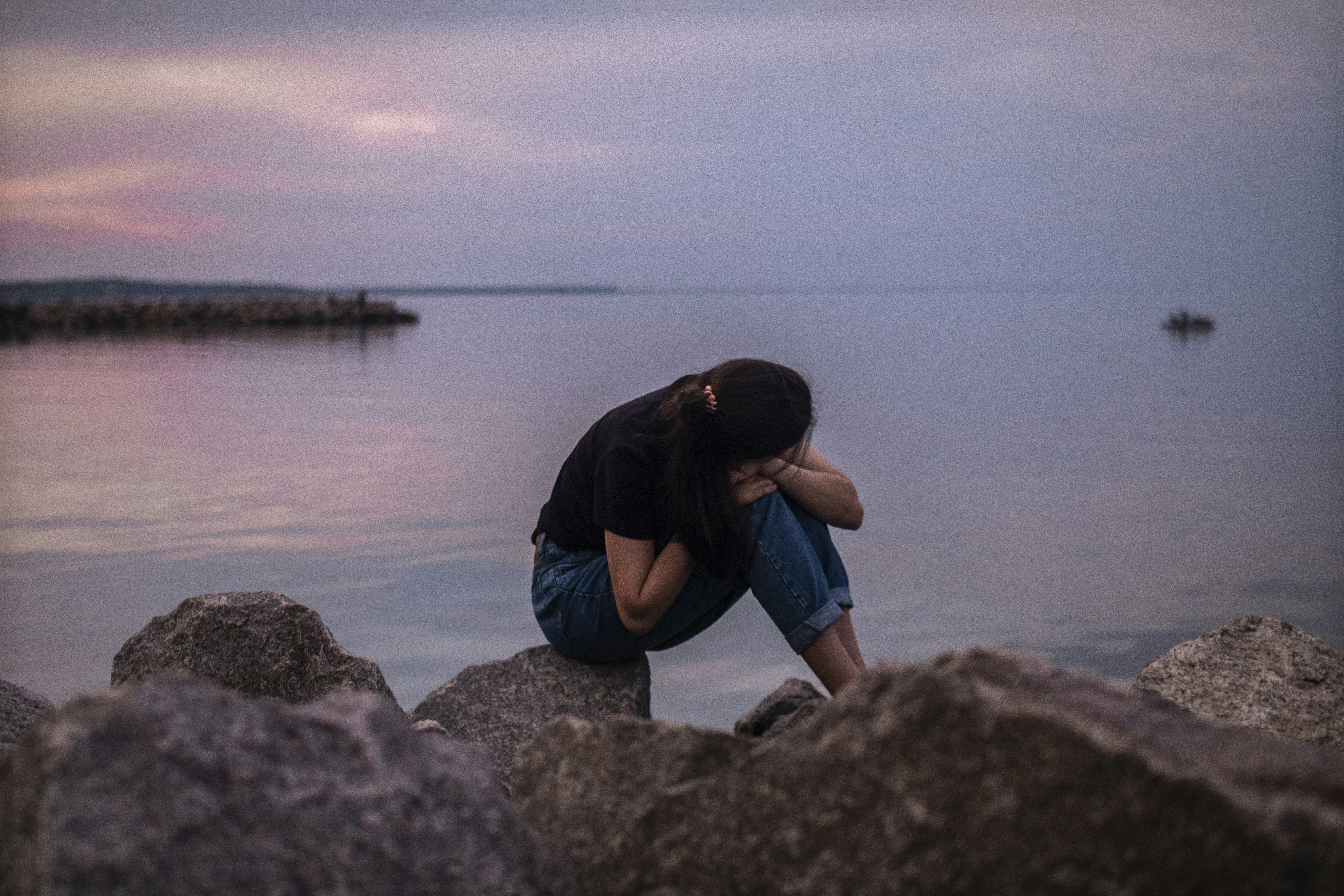 woman in black jacket and blue denim jeans sitting on rock near body of water during