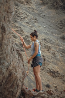 A person is climbing a steep rocky surface, wearing a sleeveless shirt and shorts. They are holding a bright orange climbing rope and appear to be preparing or adjusting their equipment. The background features rugged, rocky terrain.