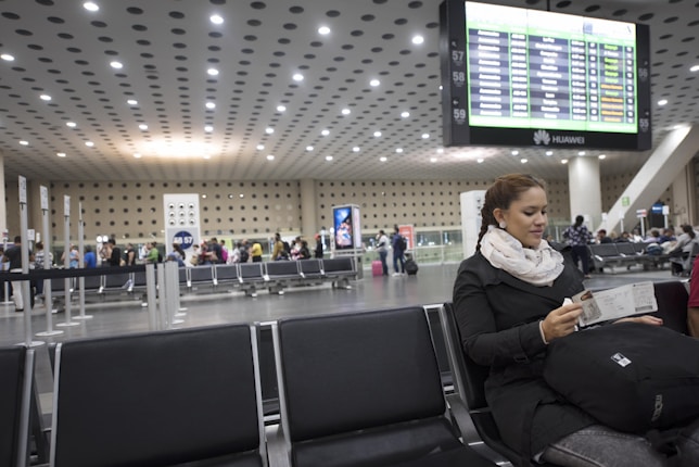 A woman with braided hair sits in a modern airport terminal, holding travel documents. She is dressed in a dark coat with a large scarf. The terminal features rows of empty black seating and a crowd gathered near check-in counters. A large digital flight information display board is visible in the background.