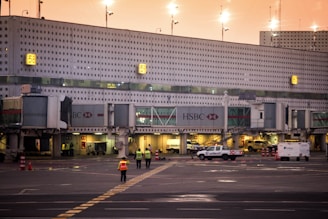 The image captures an airport terminal with multiple gates visible, marked by numbers. Jet bridges and service vehicles can be seen, along with workers wearing high-visibility vests walking on the tarmac. The lighting suggests it is either early morning or late evening, with a warm, orange hue in the sky.