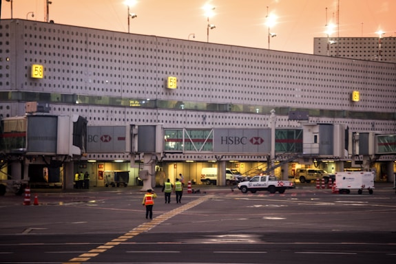 The image captures an airport terminal with multiple gates visible, marked by numbers. Jet bridges and service vehicles can be seen, along with workers wearing high-visibility vests walking on the tarmac. The lighting suggests it is either early morning or late evening, with a warm, orange hue in the sky.