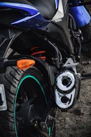 A close-up shot of a motorcycle's rear section featuring the suspension coil with a bright orange color, a visible exhaust pipe with a chrome finish, and a rugged tire with a green strip. The body is sleek, with some blue panels and a brand logo visible.