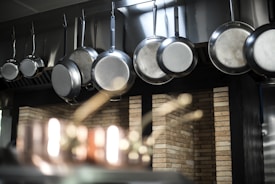 A row of shiny metal frying pans hanging from hooks against a backdrop of brick wall tiles, with soft focus lighting and reflections accentuating the texture and material of the pans and the background.