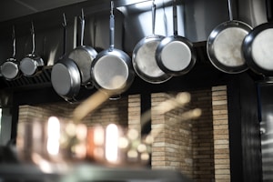 A row of shiny metal frying pans hanging from hooks against a backdrop of brick wall tiles, with soft focus lighting and reflections accentuating the texture and material of the pans and the background.