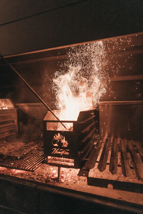 A flammaease silicone brush resting beside a hot grill with glowing coals in the background.