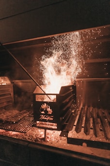 A glowing fire is burning within a metal grill setup, with sparks flying upward. A tool is resting on the grill, which has the word 'Flamfires' etched on its surface.