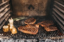 Various cuts of meat and corn are being grilled on a barbecue. The grill marks are visible on the meats, and there is a smoky ambiance. Corn on the cob and a piece of leek are also present on the grill, adding to the rustic outdoor cooking setting.