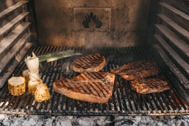 Various cuts of meat and corn are being grilled on a barbecue. The grill marks are visible on the meats, and there is a smoky ambiance. Corn on the cob and a piece of leek are also present on the grill, adding to the rustic outdoor cooking setting.