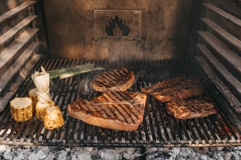 Various cuts of meat and corn are being grilled on a barbecue. The grill marks are visible on the meats, and there is a smoky ambiance. Corn on the cob and a piece of leek are also present on the grill, adding to the rustic outdoor cooking setting.