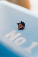 A pin featuring an illustrated face of a man wearing a baseball cap is placed on a surface with the partial text 'No 1'. The surface is light blue, and the pin contrasts sharply with it due to its darker colors.