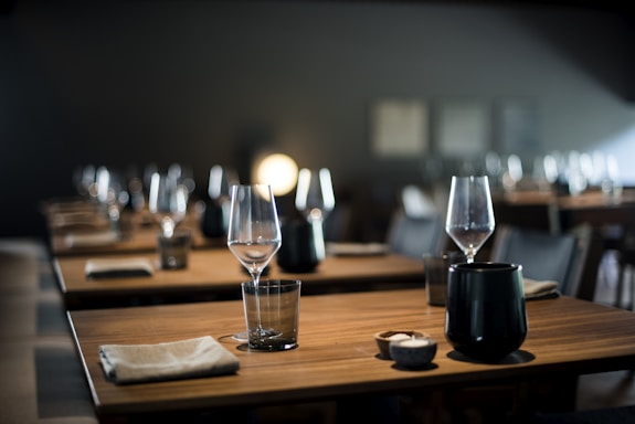 A dimly lit restaurant setting featuring neatly arranged tables with wine glasses, small black ceramic dishes, and folded cloth napkins. The atmosphere appears calm and elegant, with a soft, warm light illuminating the scene.