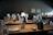 A dimly lit restaurant setting featuring neatly arranged tables with wine glasses, small black ceramic dishes, and folded cloth napkins. The atmosphere appears calm and elegant, with a soft, warm light illuminating the scene.