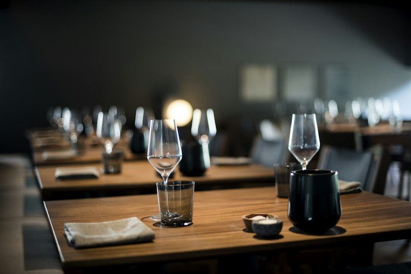 A dimly lit restaurant setting featuring neatly arranged tables with wine glasses, small black ceramic dishes, and folded cloth napkins. The atmosphere appears calm and elegant, with a soft, warm light illuminating the scene.