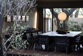 A modern dining room featuring a large rectangular table surrounded by dark chairs. A round, glowing pendant light hangs above the table, adding a warm ambiance to the room. The space is complemented by a natural setting visible through large windows, with trees and greenery creating a serene atmosphere. Decorative wall lights and a console table with various items further enhance the room's stylish appearance.