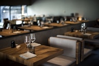 Tables set with wine glasses and cloth napkins inside a dimly lit, modern restaurant featuring wooden furniture and soft lighting.