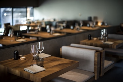 Tables set with wine glasses and cloth napkins inside a dimly lit, modern restaurant featuring wooden furniture and soft lighting.
