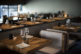 Tables set with wine glasses and cloth napkins inside a dimly lit, modern restaurant featuring wooden furniture and soft lighting.