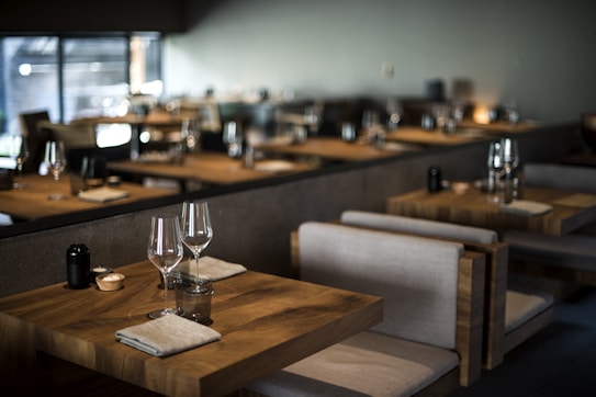 Tables set with wine glasses and cloth napkins inside a dimly lit, modern restaurant featuring wooden furniture and soft lighting.