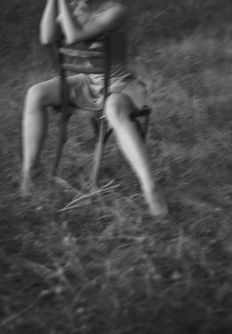 A soft-focus black and white image captures a person sitting on a wooden chair in a grassy field. The person is holding their head with their hands, and their legs are slightly apart, giving a relaxed yet introspective posture.