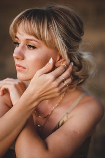 A woman with blonde hair styled in loose curls gazes thoughtfully to the side. Her makeup includes winged eyeliner and neutral-toned eyeshadow. She is adorned with several gold rings and a delicate gold chain necklace, and her manicure is a light, natural shade. The lighting casts a warm, golden hue, highlighting her facial features.