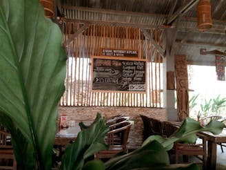 Wooden chairs and tables are arranged in an open-air dining space, accentuated by large green leaves in the foreground. A chalkboard sign with various menu items and the quote 'A goal without a plan is just a wish' is visible against vertical wooden slats. The setting appears rustic with elements like stone and wooden beams contributing to a cozy atmosphere.