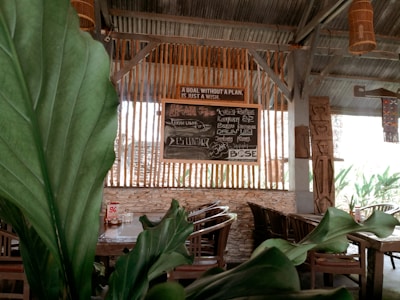 Wooden chairs and tables are arranged in an open-air dining space, accentuated by large green leaves in the foreground. A chalkboard sign with various menu items and the quote 'A goal without a plan is just a wish' is visible against vertical wooden slats. The setting appears rustic with elements like stone and wooden beams contributing to a cozy atmosphere.