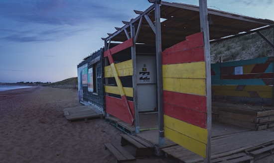 A rustic beach shack made with colorful wooden planks, featuring red, yellow, and black designs. It is situated on a sandy beach with an overcast evening sky. The structure appears homemade, constructed with wooden pallets, and sits near dunes covered with grass.