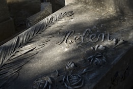 An engraved stone surface features floral carvings and a stylized leaf pattern alongside the name Helen. Shadows fall across the textured surface, giving it a somber atmosphere.